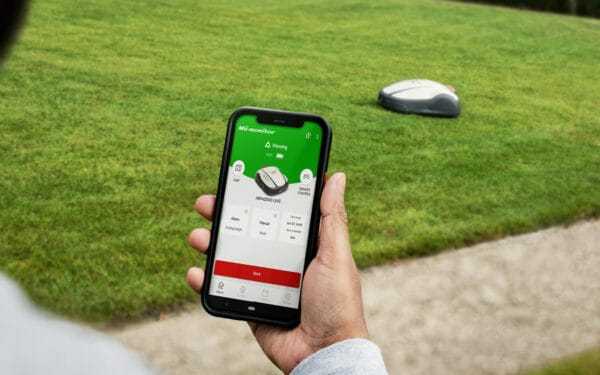 En hand håller en mobil där man ser inuti en app hur man sköter Honda Miimo robotgräsklipparen som åker i bakgrunden på en grön gräsmatta