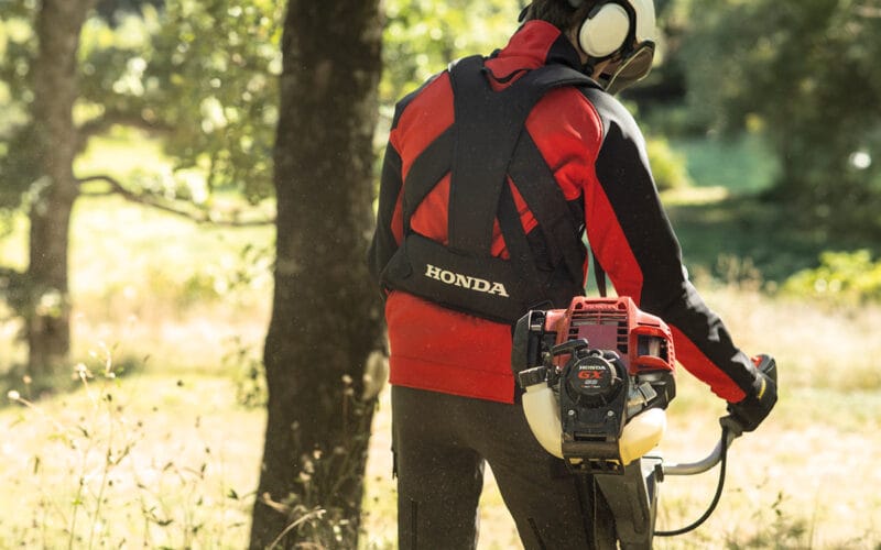 En man på en grön äng med en röjsåg från Honda i händerna. Med träd i bakgrunden