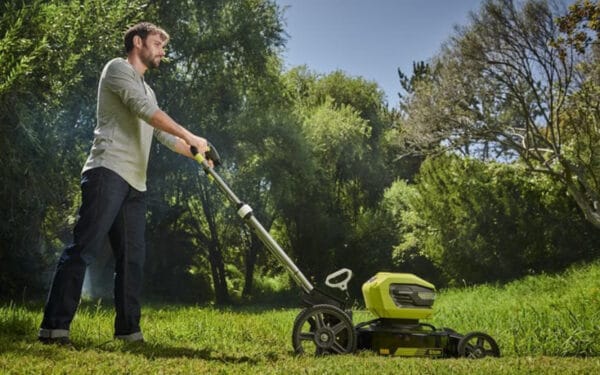 En man som kör en RYOBI gräsklippare RY36LMMX51A_140 på en grön gräsmatta med gröna stora träd i bakgrunden