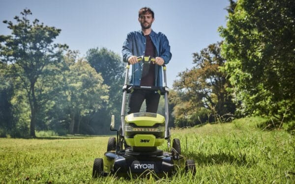 En man som kör en RYOBI gräsklippare RY36LMMX51A_140 på en grön gräsmatta med gröna stora träd i bakgrunden