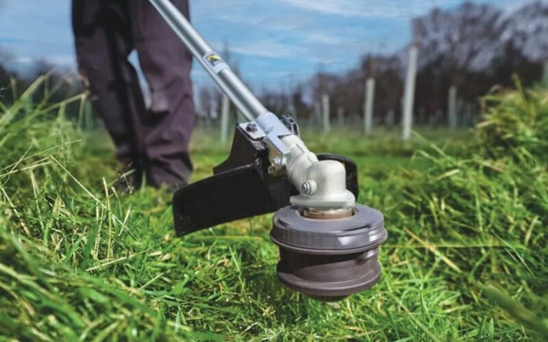En närbild på RYOBI grästrimmer RY36LTX40A-0 som klipper gräset, i bakgrunden syns två ben och träd