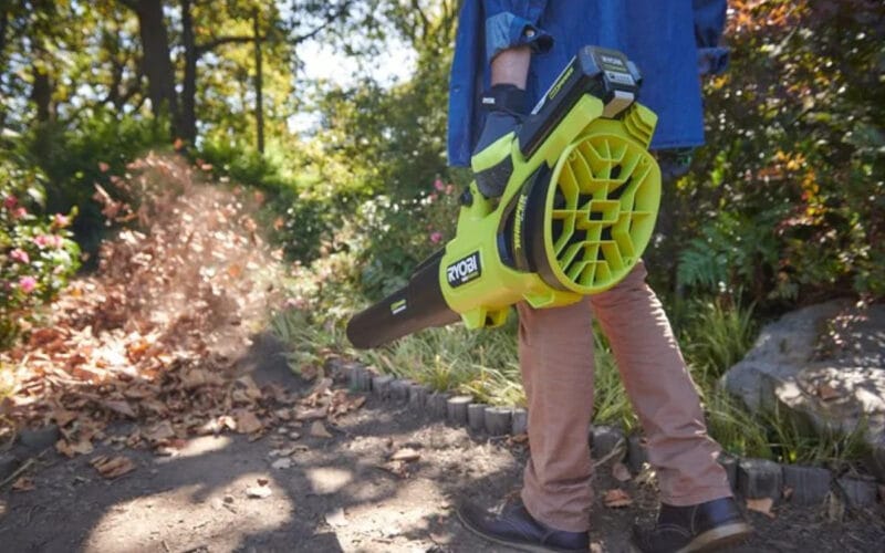 En man håller i en lövblås RY36BLXB-0 från Ryobi med löv, träd och gröna växter i bakgrunden